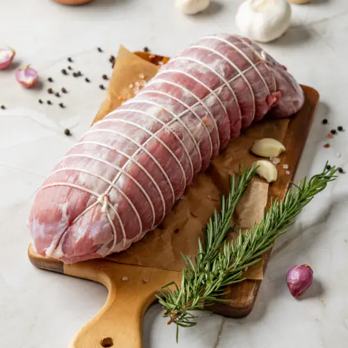 "Raw tied pork roast on wooden board with rosemary, garlic, shallots, and peppercorns prepared for cooking."